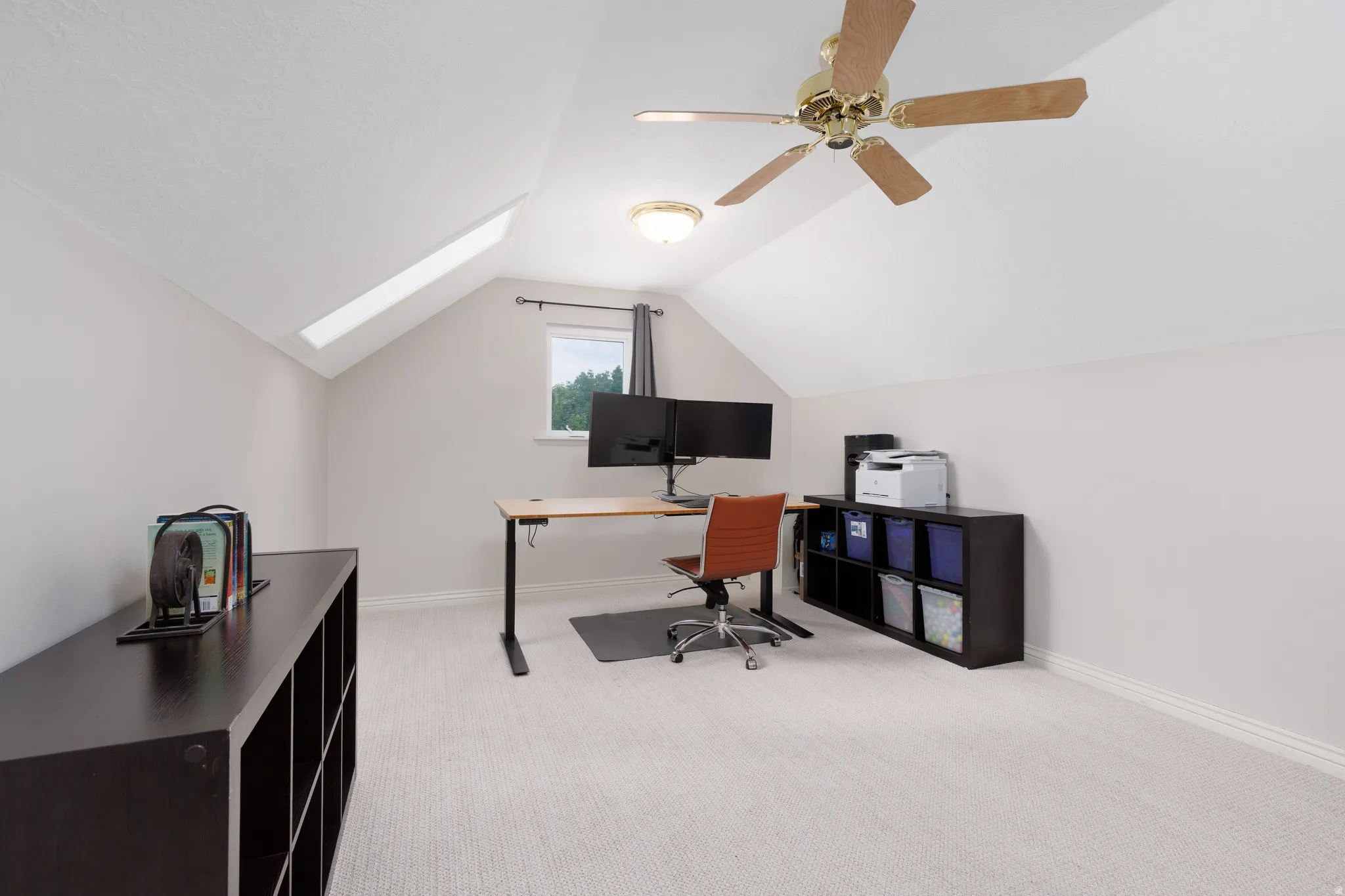 Carpeted room featuring a sloped ceiling with a skylight and window, a ceiling fan with light fixture, and white baseboards