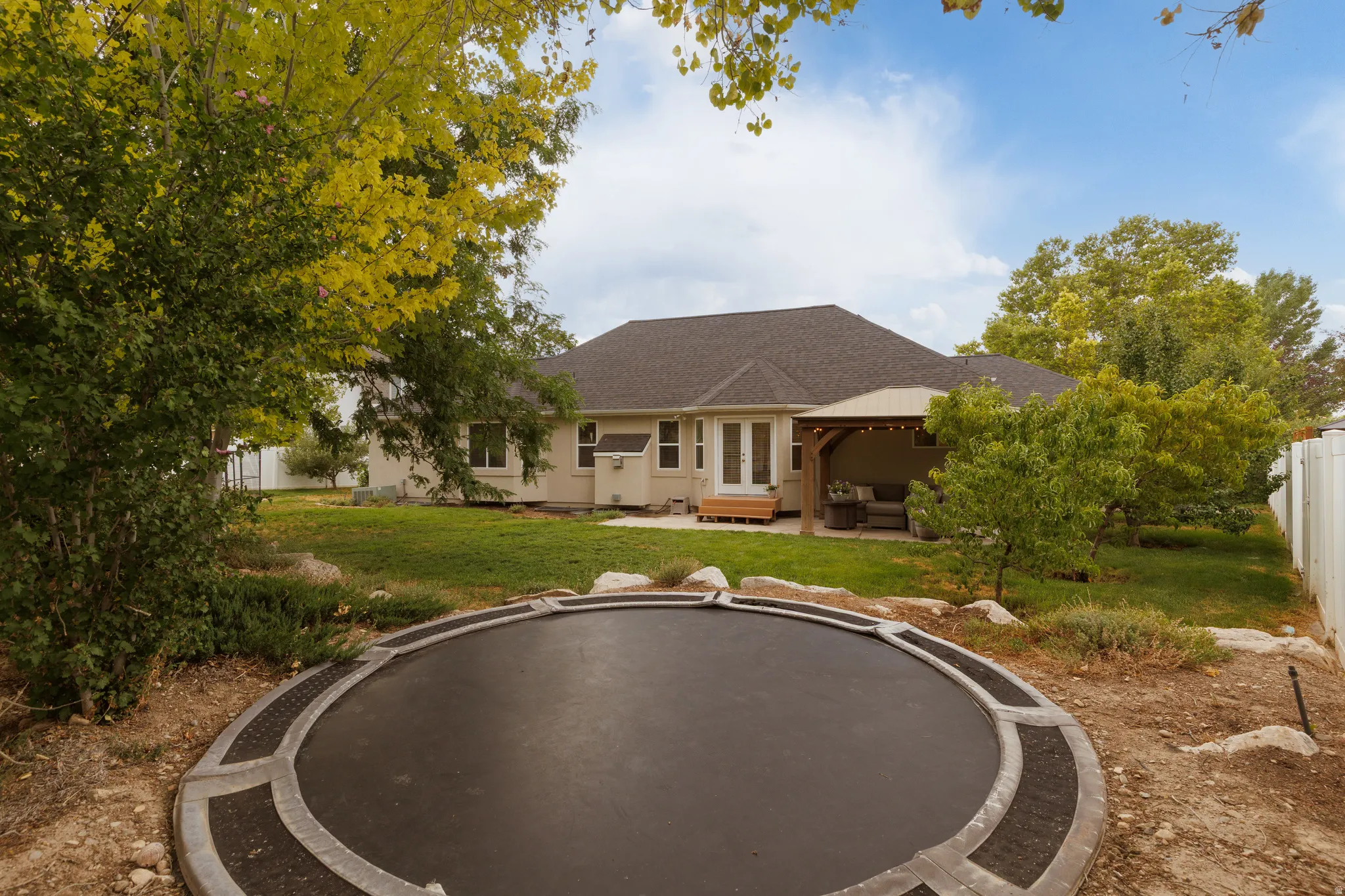Spacious backyard featuring a built-in circular ground trampoline, a covered patio with string lighting, and a well-maintained lawn