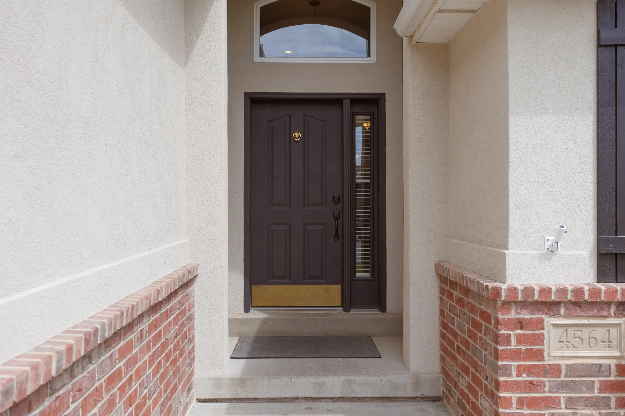 Entryway featuring a paneled front door with a transom window and sidelight, brick wainscoting, stucco exterior, and integrated address plaque