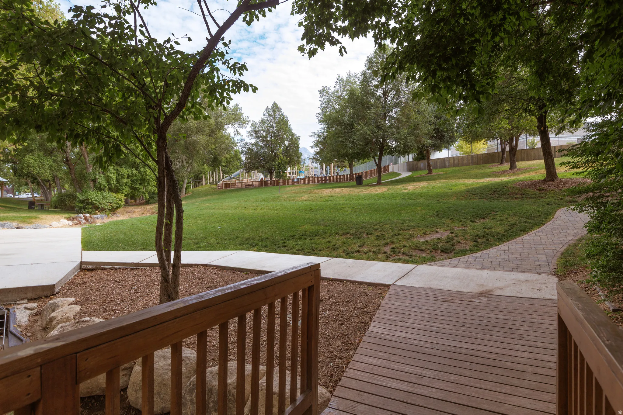 Wood-plank bridge leading to a paved pathway, expansive grassy area, and mature trees