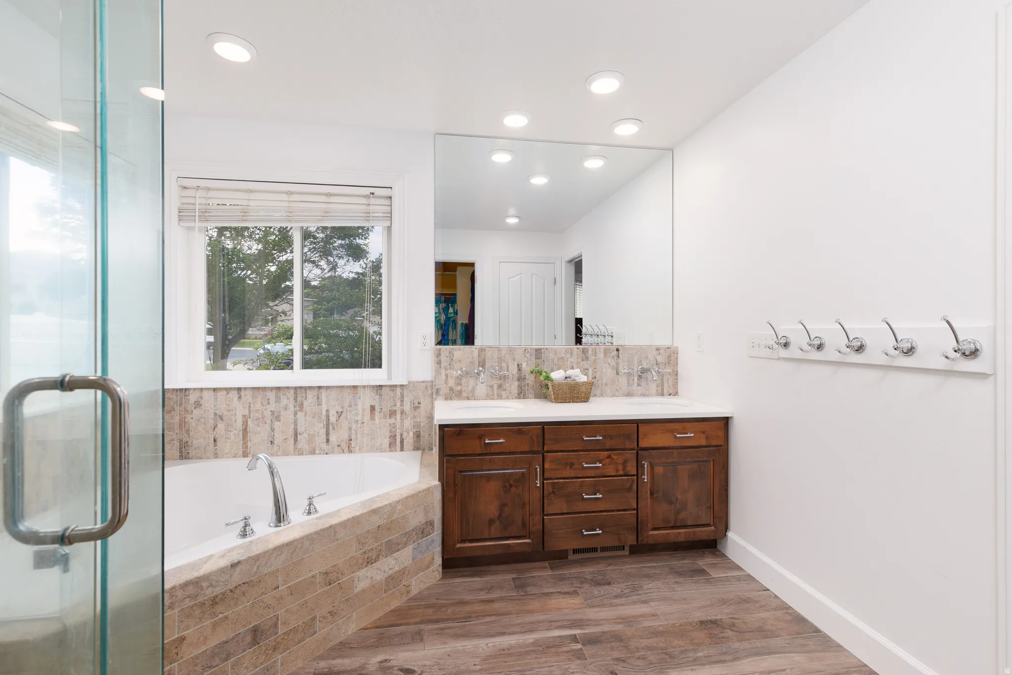 White soaking tub with tiled surround, frameless glass shower enclosure, wood-finish flooring, dark wood vanity with white countertop, and large wall-mounted mirror