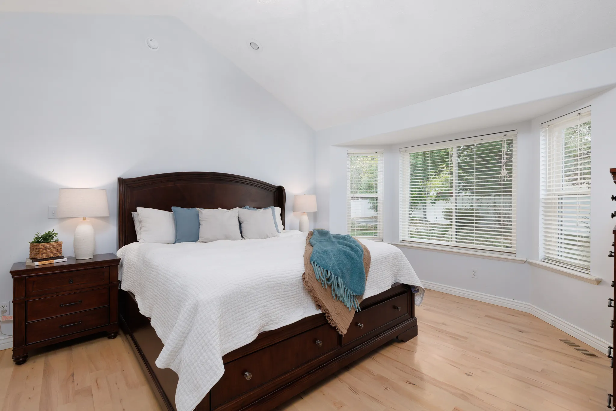 Vaulted ceiling bedroom with light wood-finish flooring, a bay window with blinds, and white painted walls