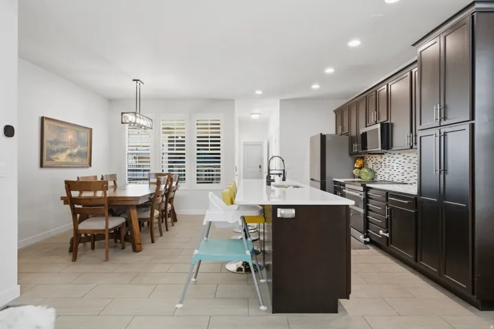 Open-concept kitchen and dining area featuring dark wood-finish cabinetry, stainless steel appliances, a white quartz island countertop, a tiled backsplash, and recessed lighting