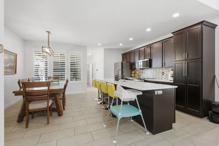 Open-concept kitchen and dining area featuring dark wood-finish cabinetry, light-toned tile flooring, a kitchen island with seating, a mosaic tile backsplash, and stainless steel appliances