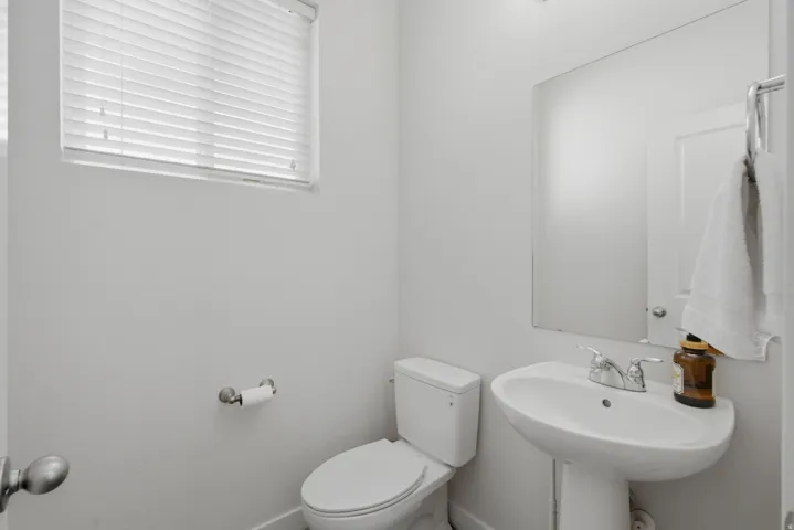 Bathroom featuring a pedestal sink with chrome fixtures, a wall-mounted mirror, a standard toilet, and a window with horizontal blinds