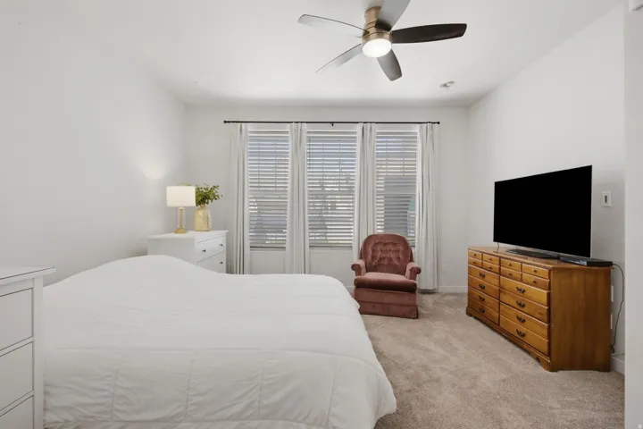 Bedroom featuring a ceiling fan, three windows with blinds and drapes, light-toned carpet flooring, and white walls