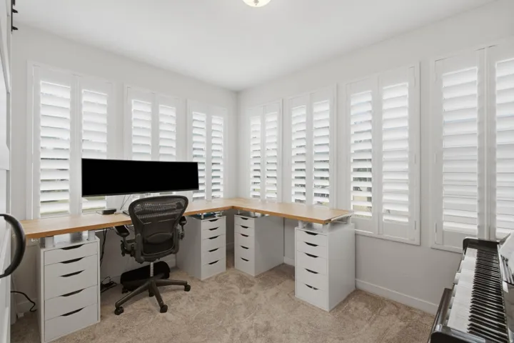 Room featuring extensive built-in desk space, multiple windows with white plantation shutters, and light-toned carpet flooring