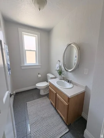 Bathroom featuring a light wood-finish vanity with a white countertop and integrated sink, a framed oval mirror, a white toilet, and gray tiled flooring