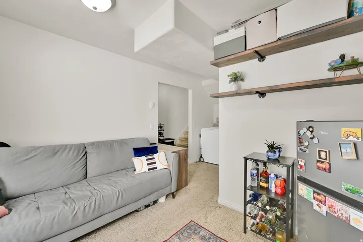 Carpeted living area with white walls, a flush-mount ceiling light, and a built-in floating shelf system featuring industrial-style pipe brackets