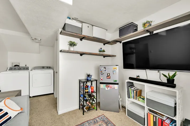 Carpeted utility area featuring a washer and dryer, adjacent to a living space with wall-mounted shelving, a built-in entertainment unit, and a compact refrigerator