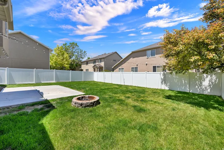 Expansive backyard featuring a manicured lawn, a large concrete patio, a built-in stone fire pit, and a full perimeter white vinyl fence