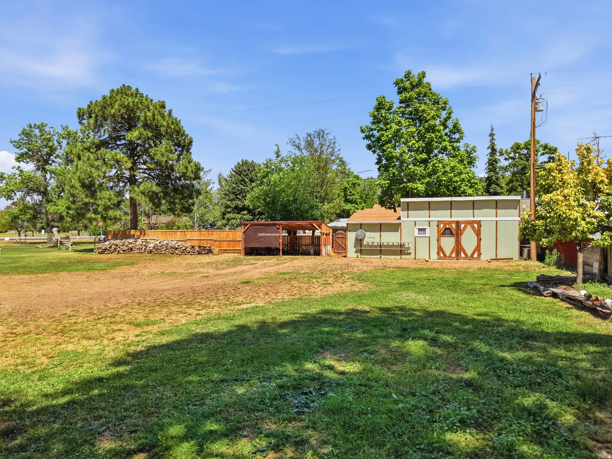 Spacious outdoor area with a light green storage building featuring wooden barn doors, a fenced enclosure, and mature trees