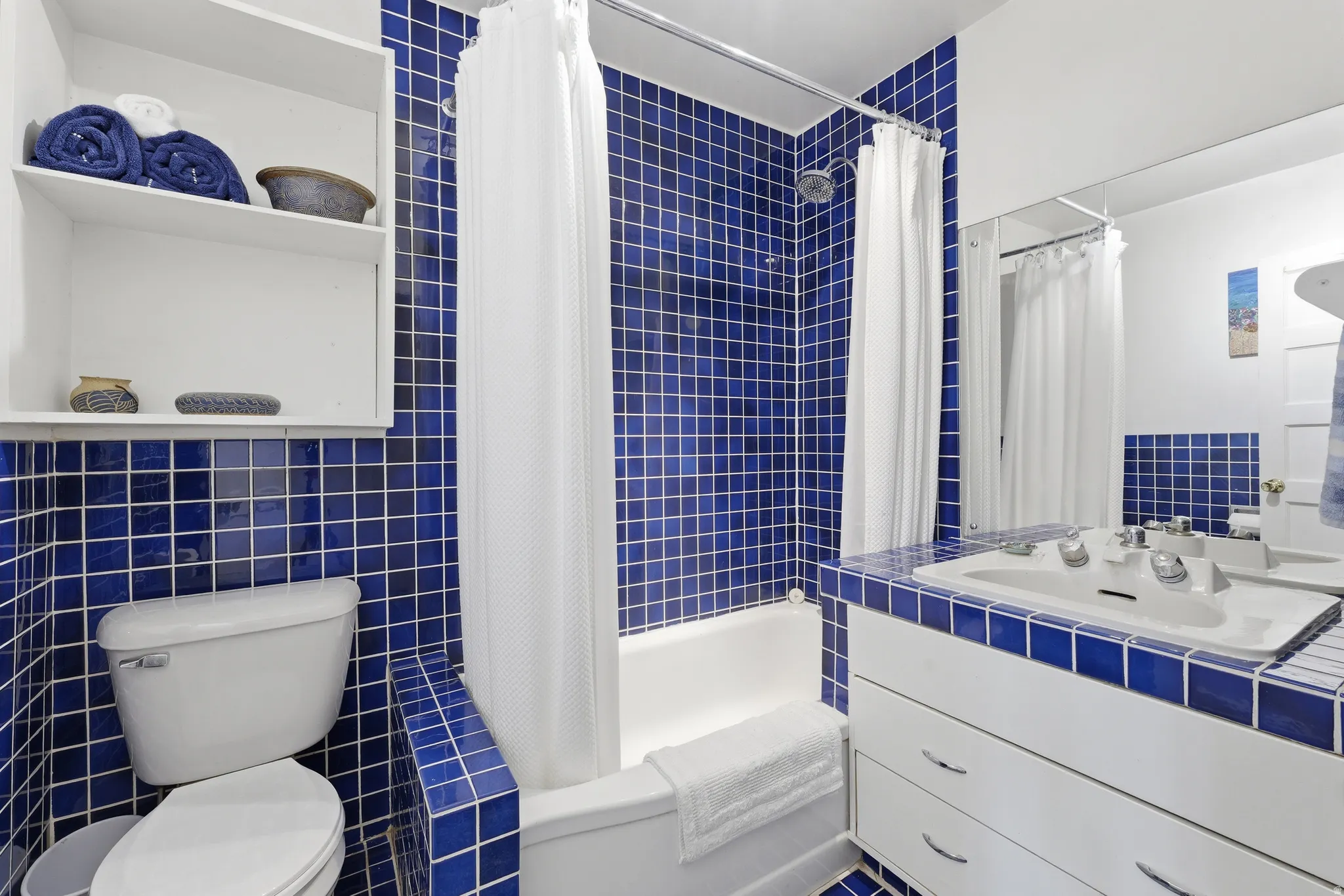 Bathroom featuring blue square wall tiling, a white bathtub with a shower, a toilet, a vanity with a drop-in sink, and a built-in mirror
