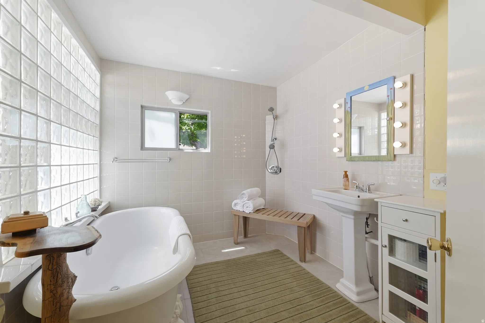 Bathroom featuring a freestanding tub, glass block wall, pedestal sink, shower with handheld spray, and a wall-mounted vanity mirror with integrated lighting