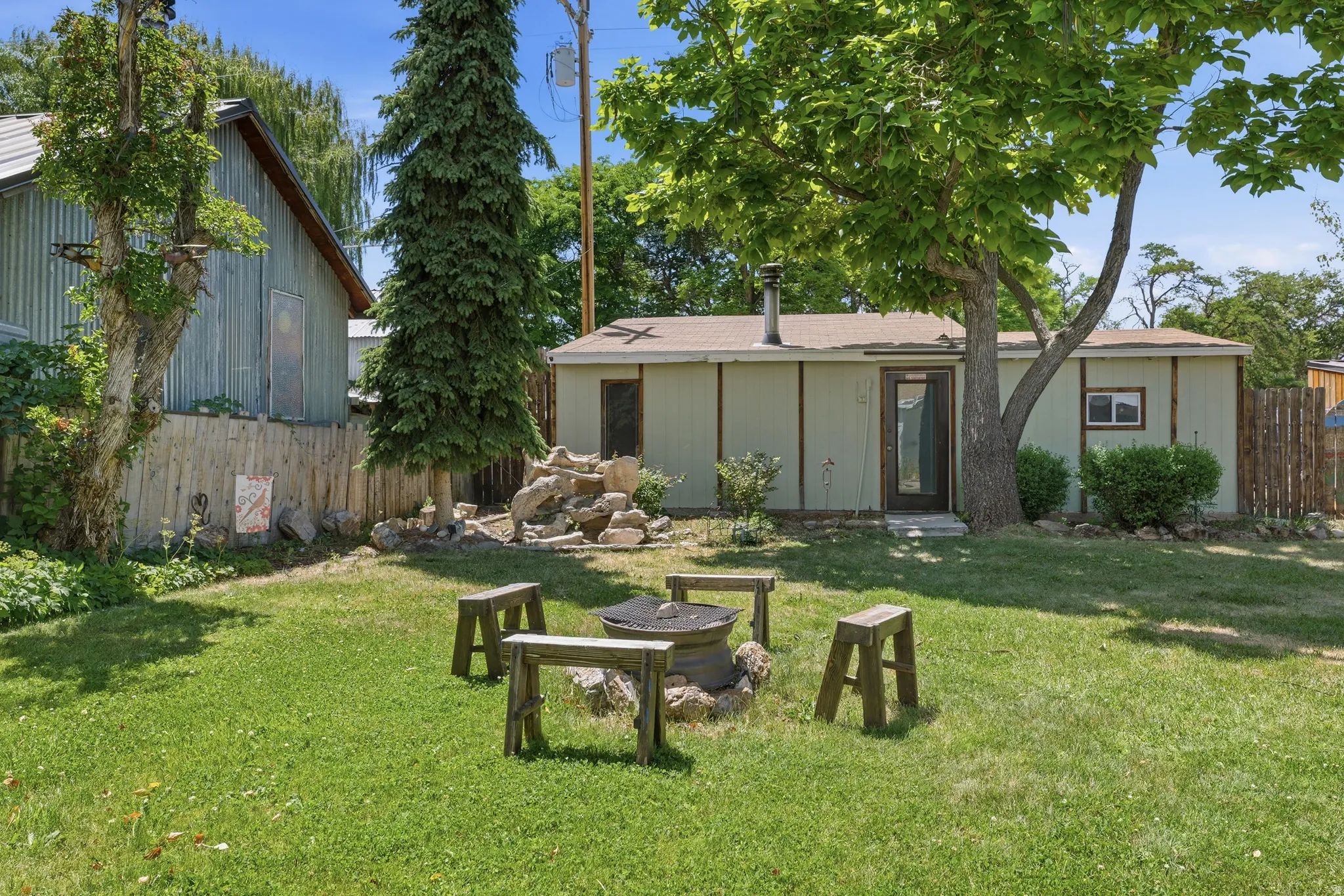 Expansive backyard featuring a lush lawn, mature trees, a built-in fire pit with log seating, a rock water feature, and a detached shed with corrugated metal siding