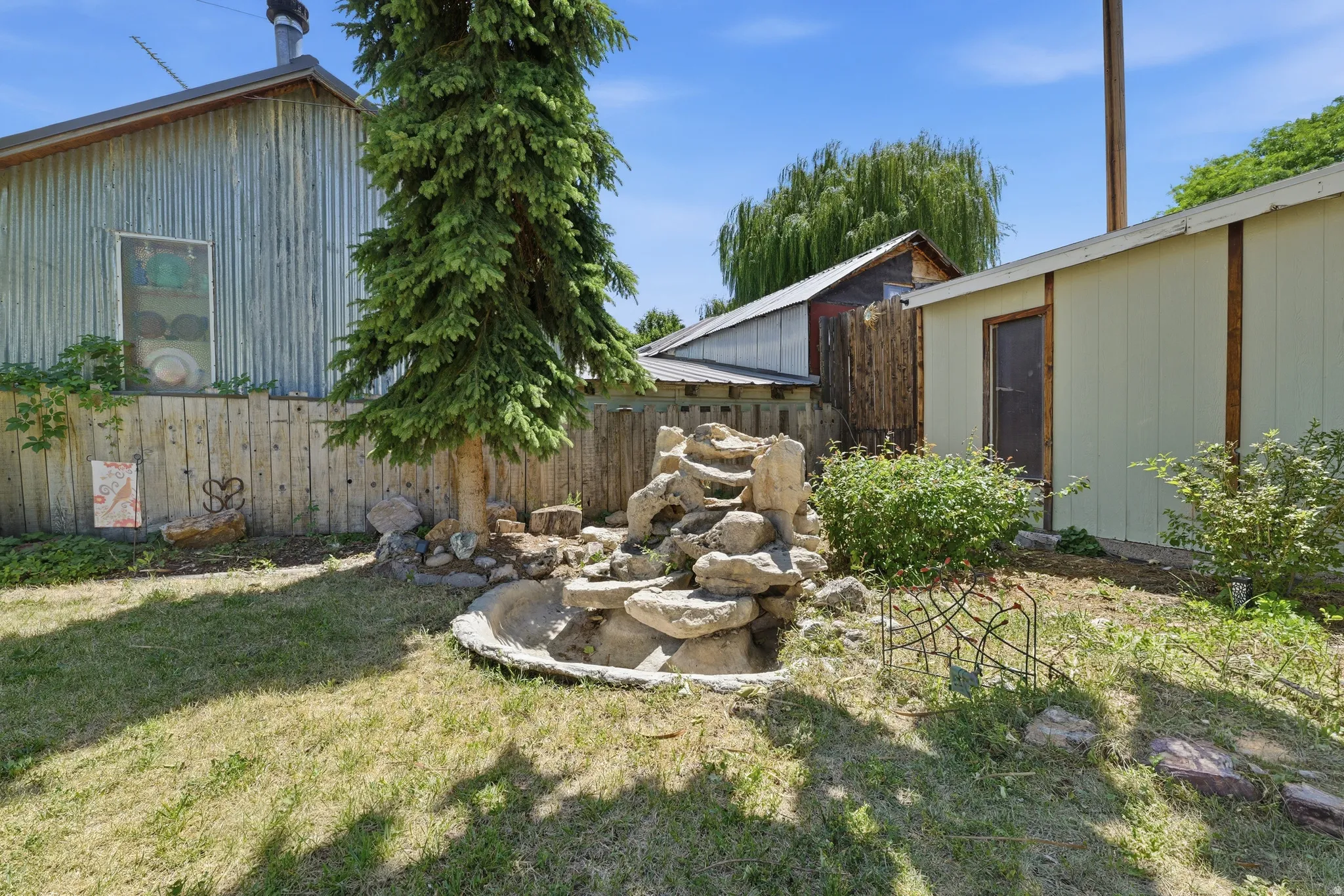 Landscaped yard featuring a tiered rock water feature, mature evergreen tree, and wood fencing