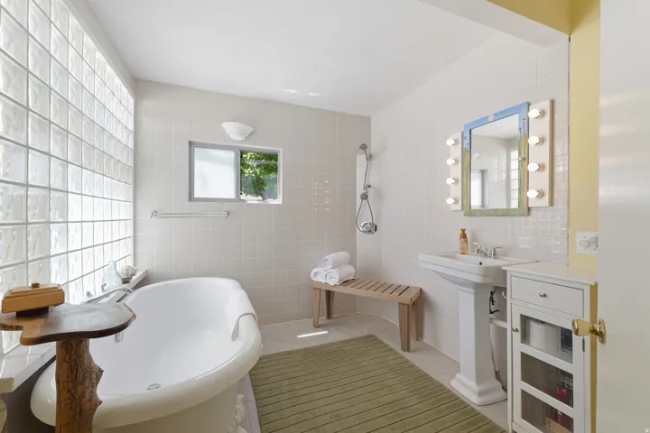 Bathroom featuring a freestanding tub, glass block wall, pedestal sink, shower with handheld spray, and a wall-mounted vanity mirror with integrated lighting