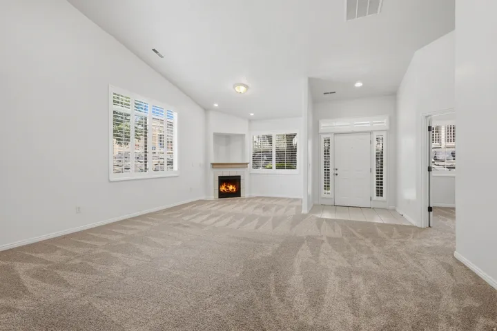 Spacious living area featuring vaulted ceilings, a fireplace with a wood mantel, large windows with plantation shutters, and light-toned carpet flooring