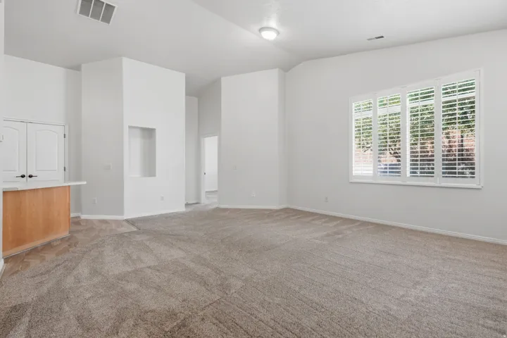 Spacious carpeted living area featuring vaulted ceilings, a large window with white plantation shutters, a recessed wall niche, and a breakfast bar with a light wood base and light-toned countertop