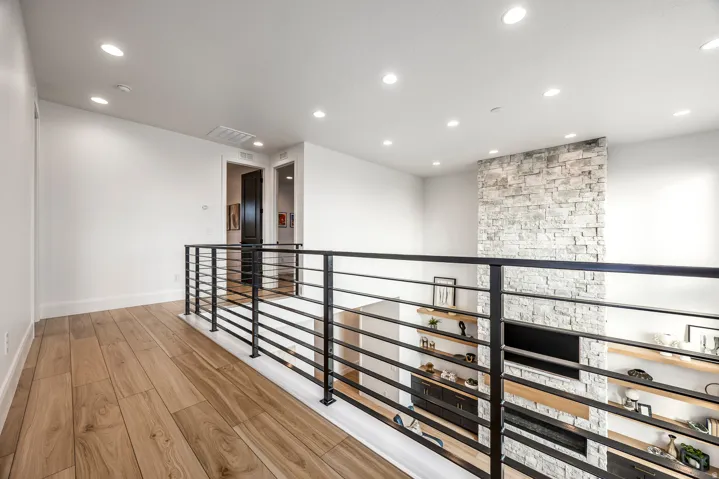 Upper-level hallway with wood-finish flooring, recessed lighting, and a contemporary black metal railing overlooking a stone-clad feature wall