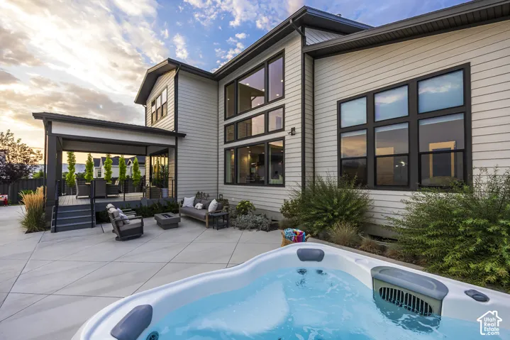 Outdoor patio featuring a hot tub, expansive concrete surfacing, a covered deck with dark railings, and a two-story exterior with large black-framed windows