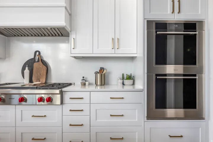 Modern kitchen featuring stainless steel appliances, stacked wall ovens, a gas range with a matching range hood, white shaker cabinetry, and light-toned countertops