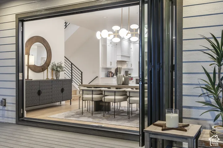 Expansive accordion-style glass doors open to reveal an open-concept interior featuring light wood-finish flooring, a modern linear chandelier, and a contemporary staircase with black metal railings