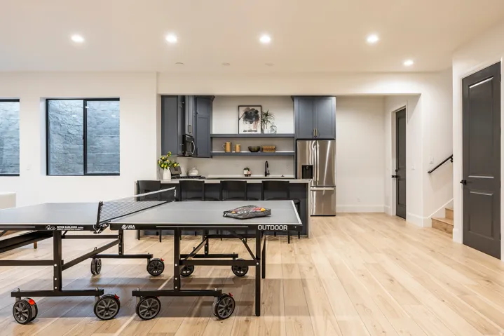 Spacious recreation area featuring light wood-finish flooring, recessed lighting, and a full wet bar with dark blue cabinetry, a stainless steel refrigerator, and built-in shelving