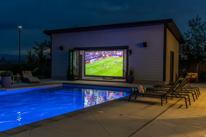 Backyard oasis featuring an illuminated swimming pool, a dedicated pool house with bifold doors, an integrated outdoor entertainment screen, and extensive concrete hardscaping