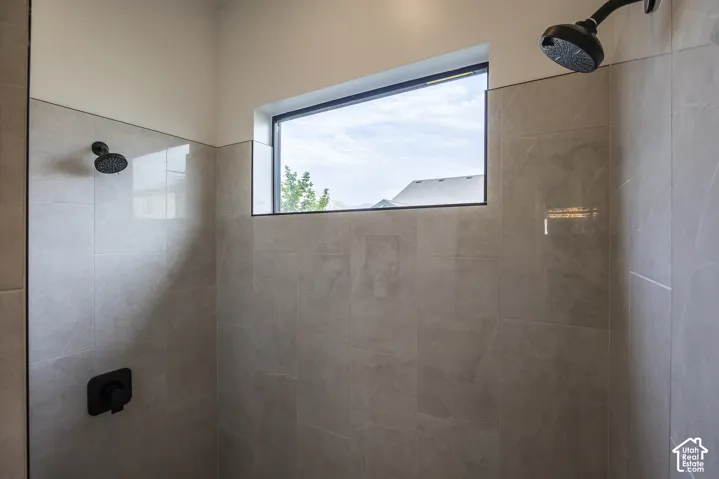 Spacious shower featuring light-toned tile walls, a horizontal window with black trim, and dual matte black shower heads
