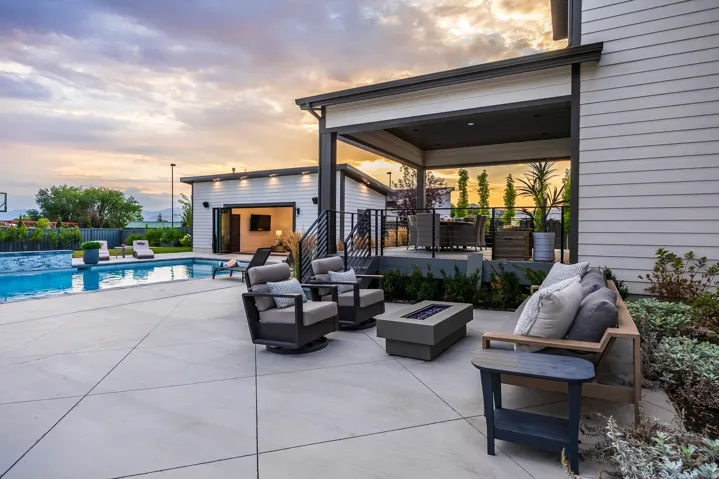 Expansive concrete patio featuring a built-in swimming pool with a stone-finish edge, a covered outdoor living area with a dark ceiling, and a separate structure with bi-fold doors