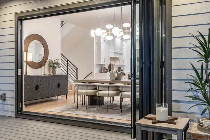 Expansive accordion-style glass doors open to reveal an open-concept interior featuring light wood-finish flooring, a modern linear chandelier, and a contemporary staircase with black metal railings