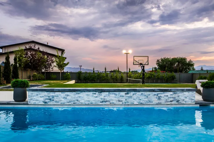 Expansive backyard featuring a sparkling swimming pool with a stone-clad retaining wall, a manicured lawn, a half-basketball court with lighting, and mature landscaping providing privacy