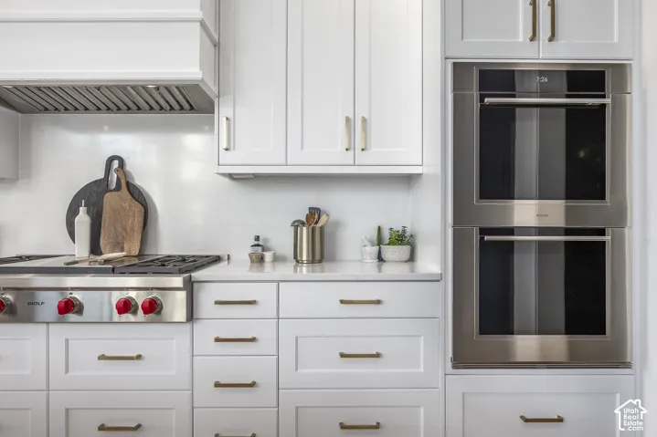 Modern kitchen featuring stainless steel appliances, stacked wall ovens, a gas range with a matching range hood, white shaker cabinetry, and light-toned countertops