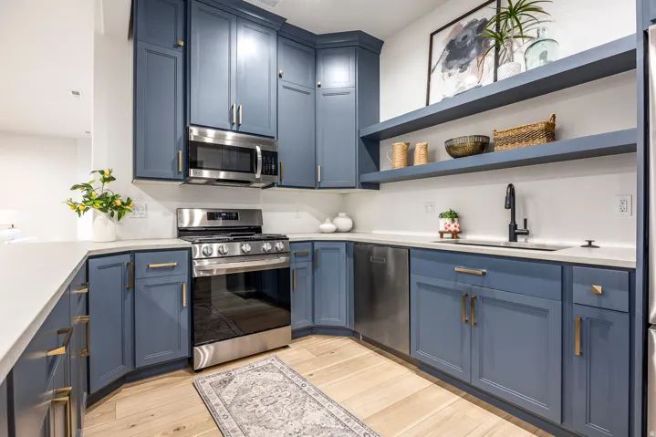 Modern kitchen featuring blue shaker-style cabinetry with gold hardware, stainless steel appliances, light-toned countertops, and wood-finish flooring