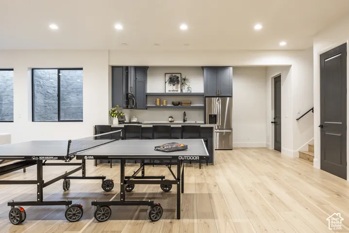 Spacious recreation area featuring light wood-finish flooring, recessed lighting, and a full wet bar with dark blue cabinetry, a stainless steel refrigerator, and built-in shelving