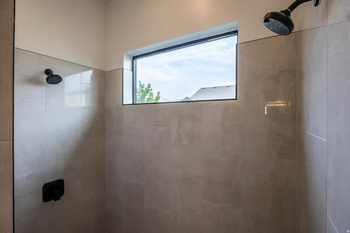 Spacious shower featuring light-toned tile walls, a horizontal window with black trim, and dual matte black shower heads