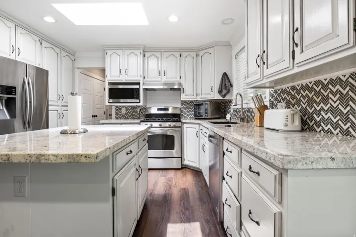 Bright kitchen featuring a central island with a light-toned countertop, extensive white cabinetry, stainless steel appliances, a stylish chevron-patterned backsplash, and rich wood-finish flooring