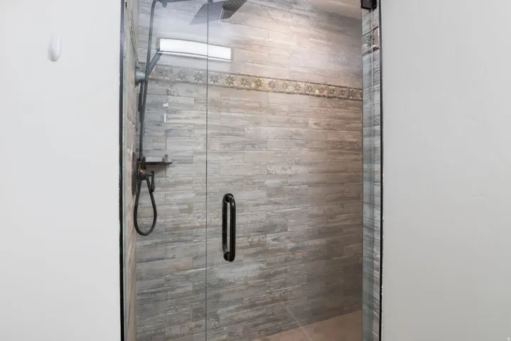Glass-enclosed shower featuring wood-look tiled walls, a decorative accent band, and dark-finish fixtures including a rain shower head and handheld sprayer