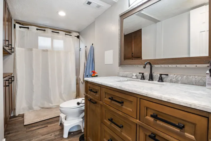Bathroom featuring wood-finish flooring, a wood vanity with dark hardware, a white countertop, and a shower with a privacy window