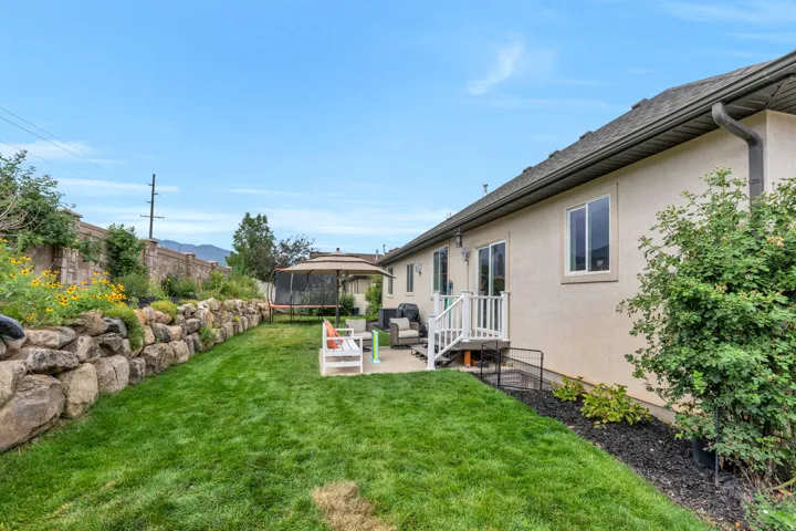 Expansive backyard featuring a stone retaining wall with plantings, a manicured lawn, a concrete patio, and a white deck with railings accessing the home