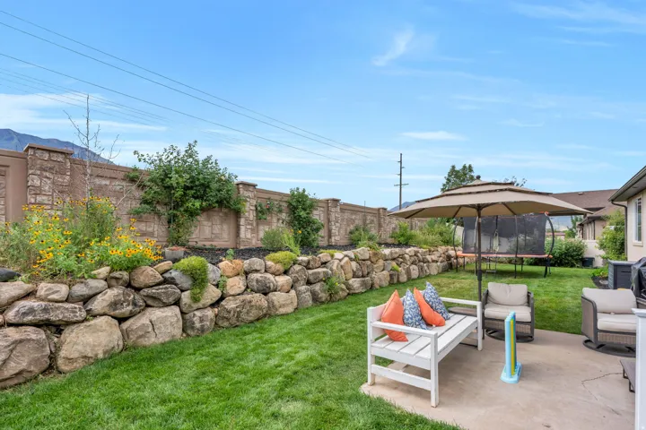 Expansive backyard featuring a stone retaining wall with mature plantings, a concrete patio area, and a stone-clad perimeter fence