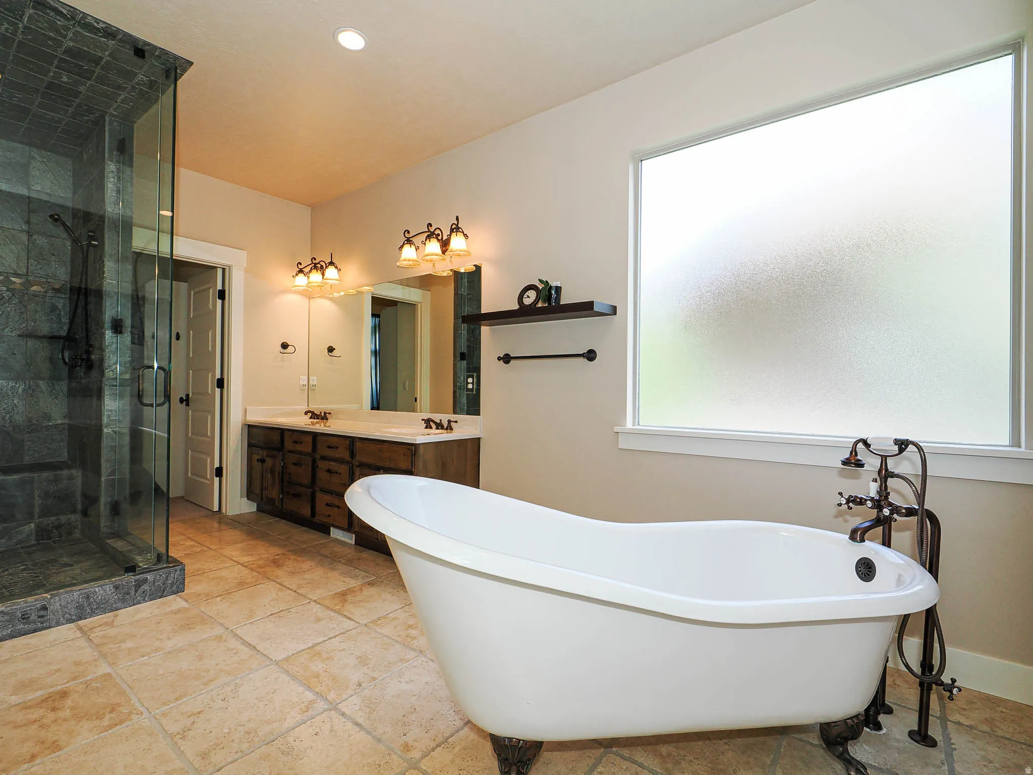 Bathroom featuring a freestanding clawfoot tub, a walk-in shower with natural stone tile, a double vanity with wood-finish cabinetry and light-colored countertops, and tiled flooring