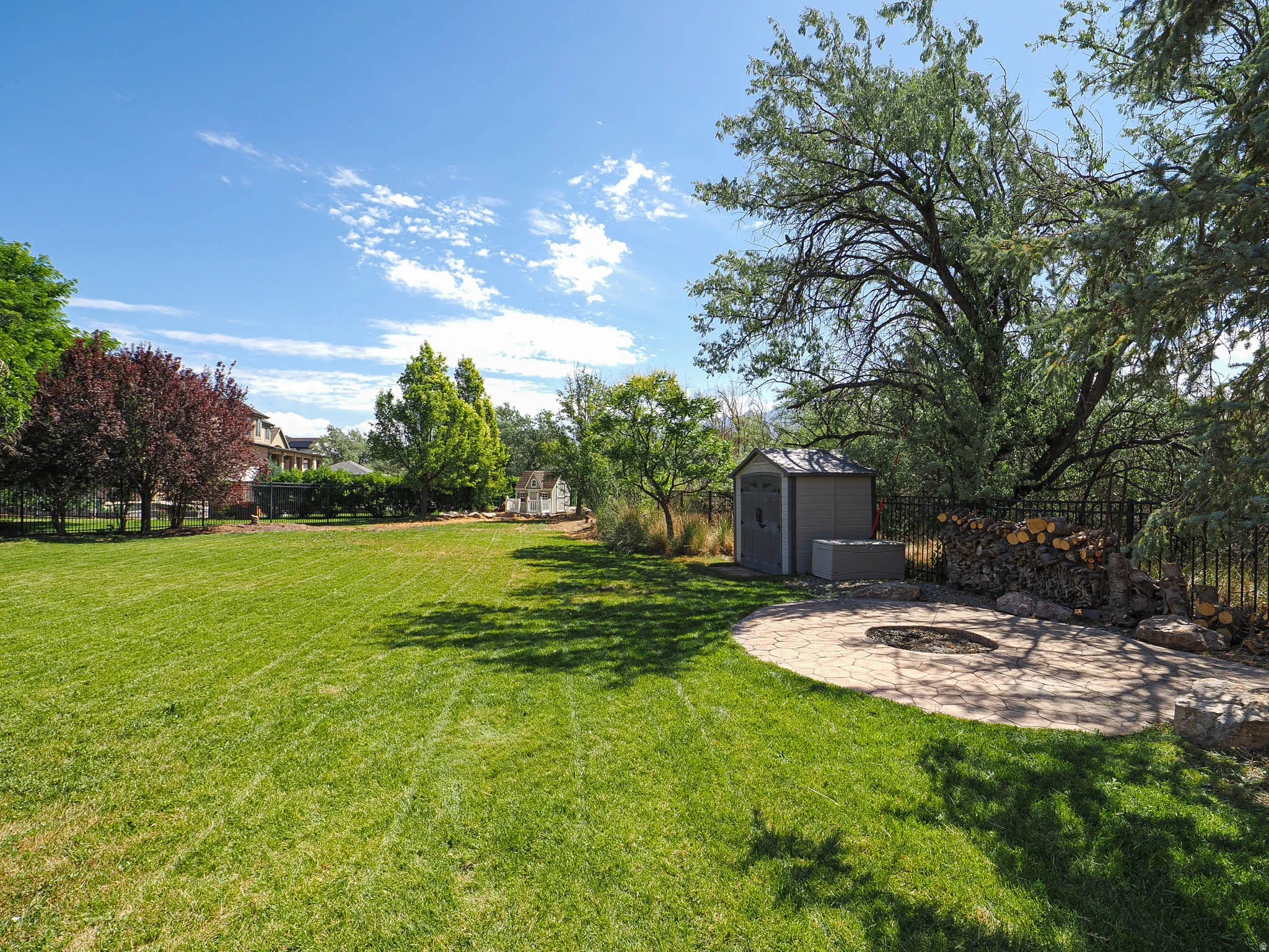 Expansive green lawn featuring a stone patio with an integrated fire pit, a storage shed, and mature trees