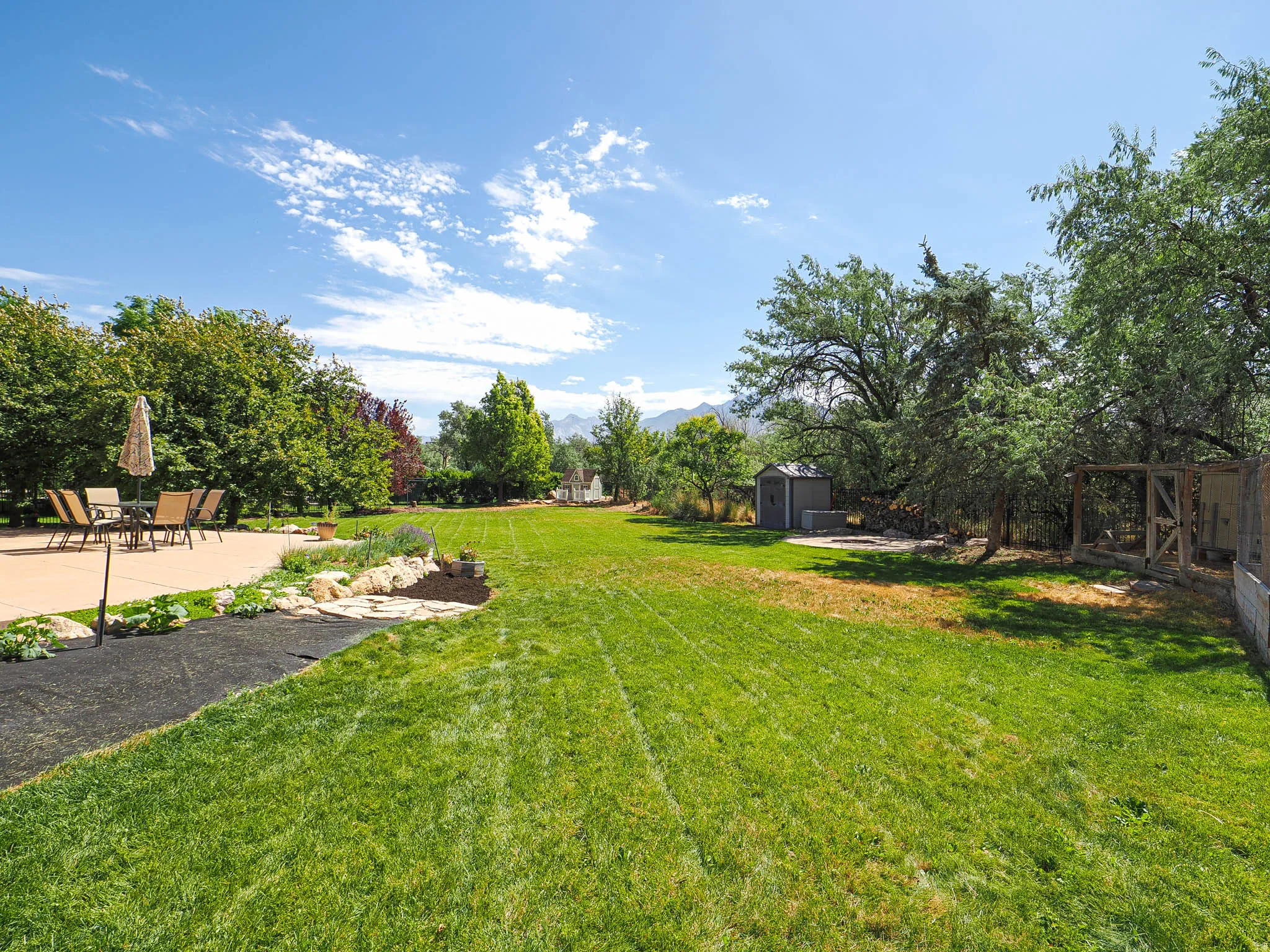 Expansive backyard featuring a large concrete patio, manicured lawn, established garden beds with rock borders, mature trees, and a utility shed