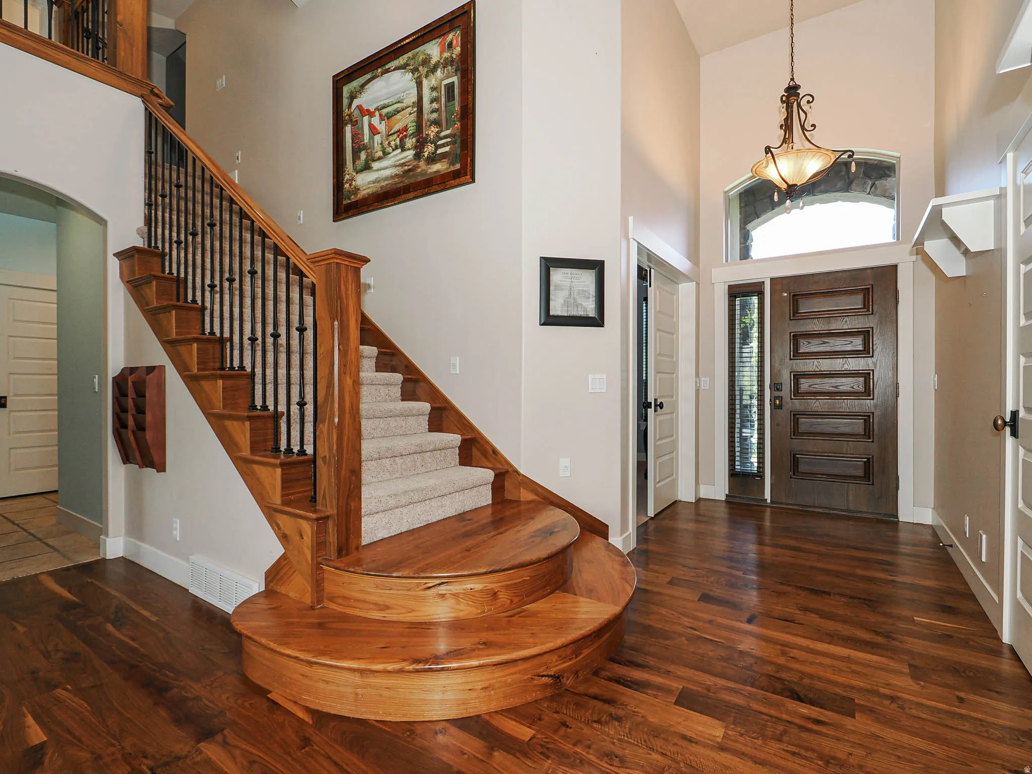 Inviting entryway featuring rich wood-finish flooring, a grand staircase with carpeted treads and wrought iron spindles, a paneled solid wood entry door with a sidelight, a transom window, and a decorative chandelier