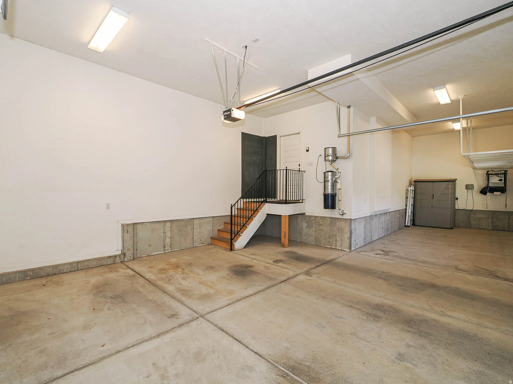 Spacious garage with concrete flooring, fluorescent overhead lighting, an interior staircase with a wrought iron railing, and an automatic garage door opener