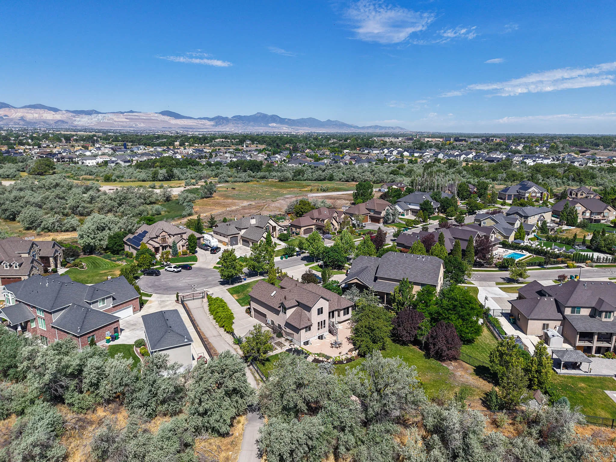 Expansive suburban landscape featuring numerous residential properties with varied architectural styles and dark shingle roofs, surrounded by mature trees and natural vegetation