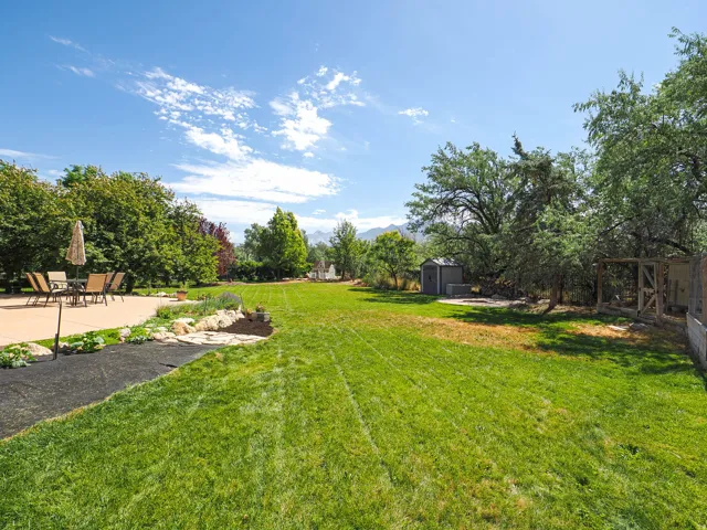 Expansive backyard featuring a large concrete patio, manicured lawn, established garden beds with rock borders, mature trees, and a utility shed