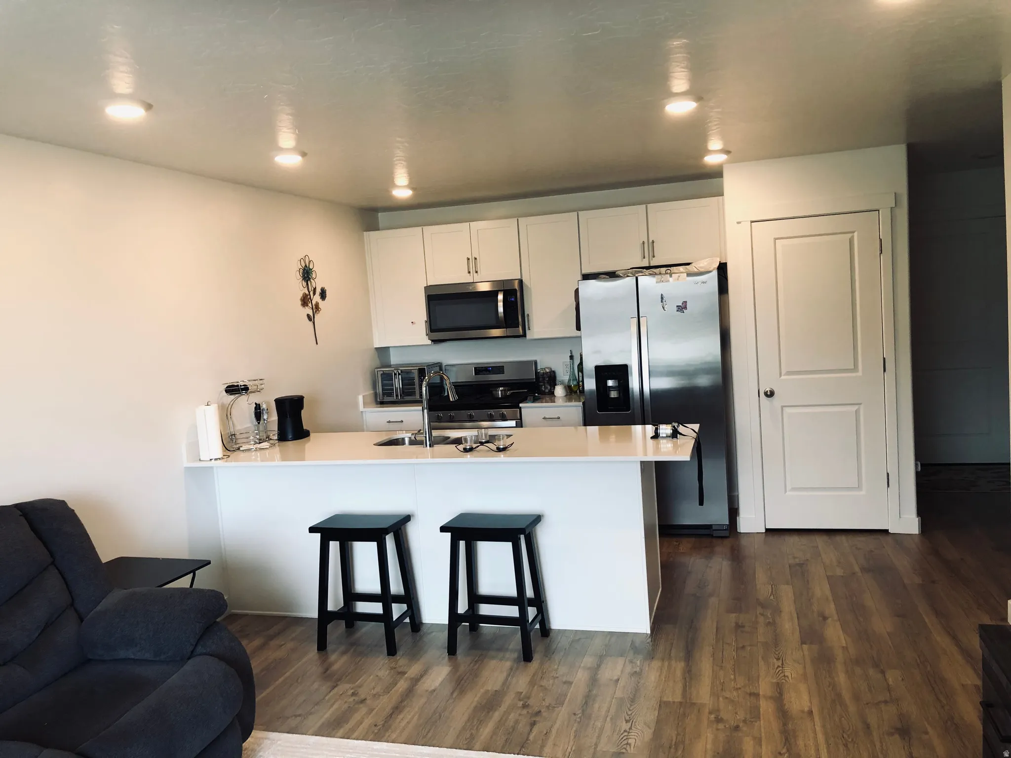 Open-concept kitchen and living area featuring white cabinetry, stainless steel appliances, a white countertop island with seating, recessed ceiling lighting, and wood-finish flooring